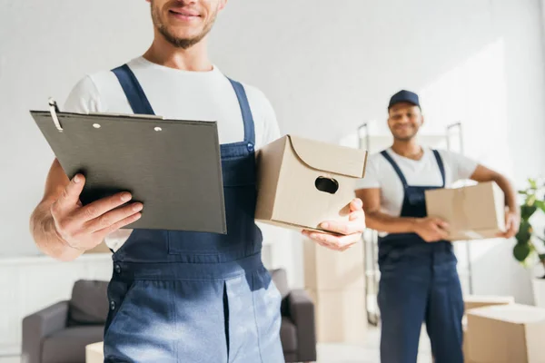 Trabajador en uniforme sujetapapeles cerca de compañero de trabajo indio con caja sobre fondo borroso - foto de stock