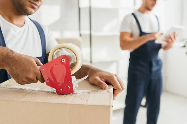 Cropped view of indian mover packing box near coworker with digital tablet on blurred background — Stock Photo