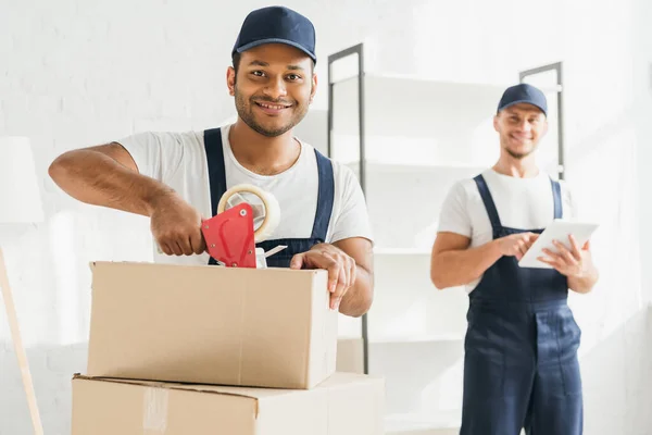 Smiling indian mover packing box near coworker with digital tablet on blurred background — Stock Photo