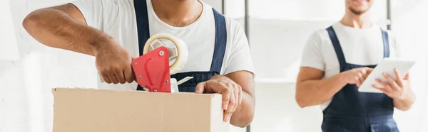 Partial view of indian mover packing box near coworker with digital tablet on blurred background, banner — Stock Photo