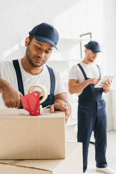 Indio mover caja de embalaje cerca de compañero de trabajo con tableta digital sobre fondo borroso - foto de stock