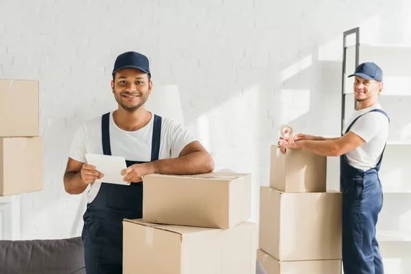 Cheerful indian mover holding digital tablet near coworker on blurred background — Stock Photo