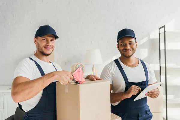 Feliz mover caja de embalaje cerca de indio compañero de trabajo utilizando tableta digital - foto de stock