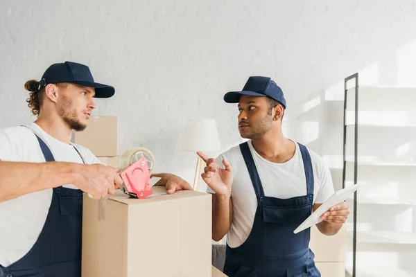 Mover caja de embalaje cerca de indio compañero de trabajo señalando con el dedo y la tableta digital - foto de stock