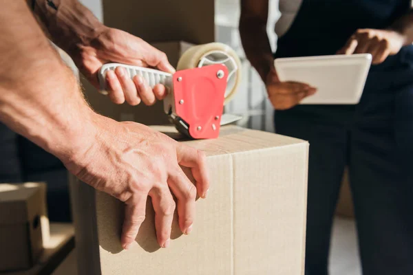 Cropped view of mover packing box with scotch tape while coworker using digital tablet — Stock Photo