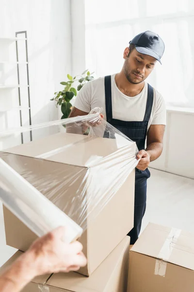 Indian mover in overalls wrapping box with coworker — Stock Photo