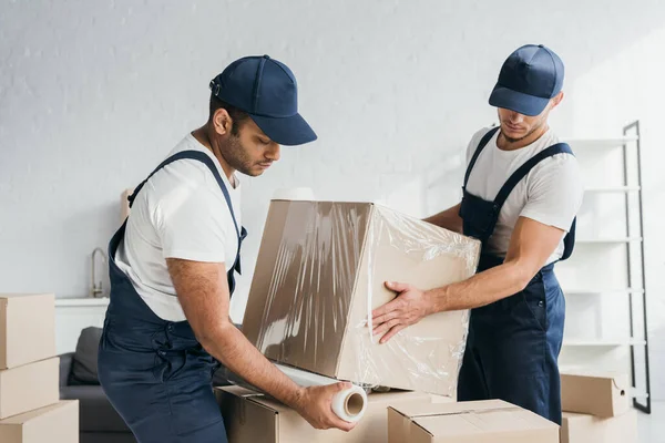 Multiethnic movers in overalls wrapping box with stretch film — Stock Photo