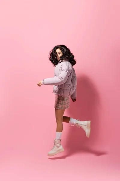 Side view of brunette young woman in stylish winter outfit jumping on pink background — Stock Photo