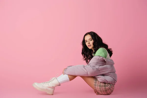 Side view of brunette young woman in stylish winter outfit posing on floor on pink background — Stock Photo