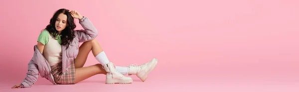 Вид збоку брюнетки молода жінка в стильному зимовому вбранні позує на підлозі на рожевому фоні, банер — стокове фото