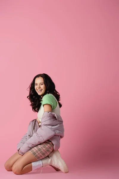 Side view of happy brunette young woman in stylish winter outfit posing on knees on pink background — Stock Photo