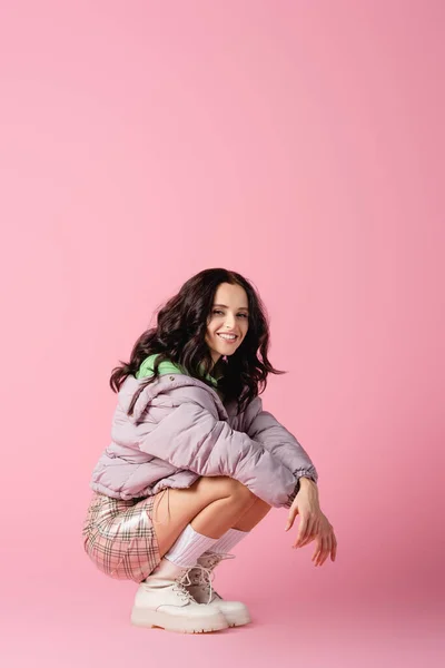 Happy brunette young woman in stylish winter outfit posing on floor on pink background — Stock Photo