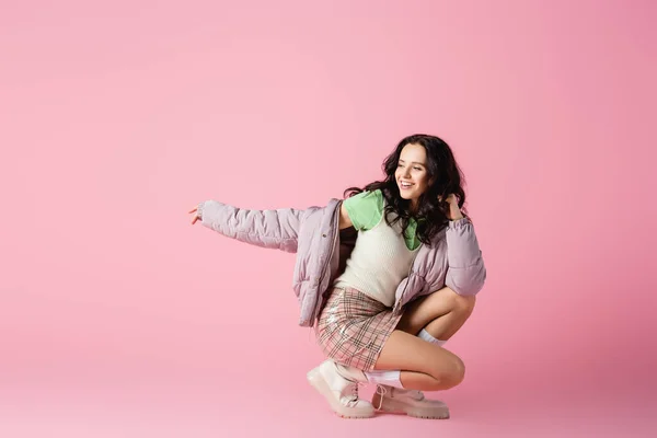 Happy brunette young woman in stylish winter outfit posing on floor on pink background — Stock Photo