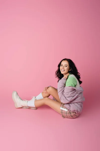 Sonriente morena joven en traje de invierno posando en el suelo sobre fondo rosa - foto de stock