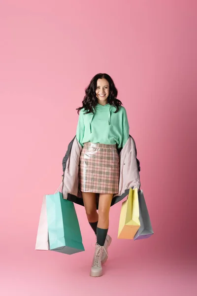 Улыбающаяся брюнетка молодая женщина в повседневном зимнем наряде с пакетами покупок на розовом фоне — стоковое фото