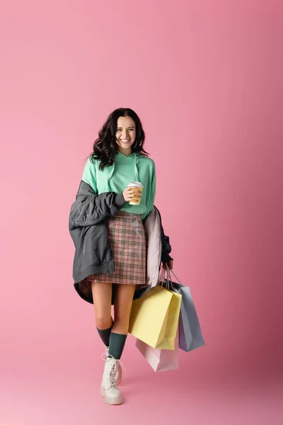 Улыбающаяся брюнетка молодая женщина в случайном зимнем наряде с пакетами для покупок и кофе, чтобы пойти на розовый фон — стоковое фото