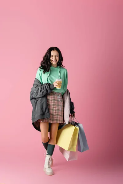 Smiling brunette young woman in casual winter outfit with shopping bags and coffee to go on pink background — Stock Photo