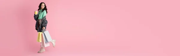 Улыбающаяся брюнетка молодая женщина в повседневной зимней одежде с пакетами для покупок и кофе, чтобы пойти на розовый фон, баннер — стоковое фото