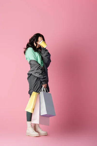 Sorrindo morena jovem em roupa de inverno casual com sacos de compras e café para ir em fundo rosa — Fotografia de Stock