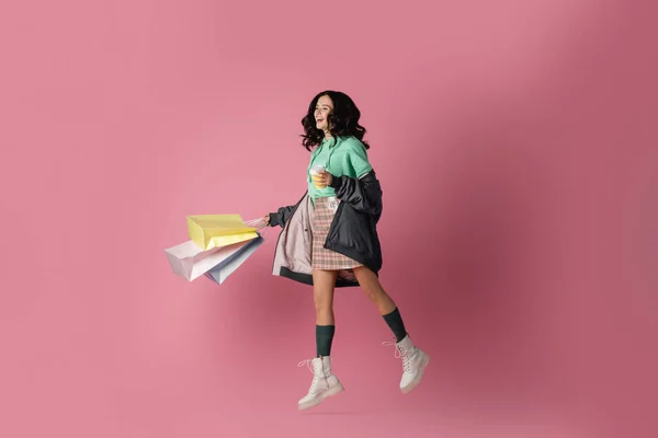 Smiling brunette young woman in casual winter outfit with shopping bags and coffee to go jumping on pink background — Stock Photo