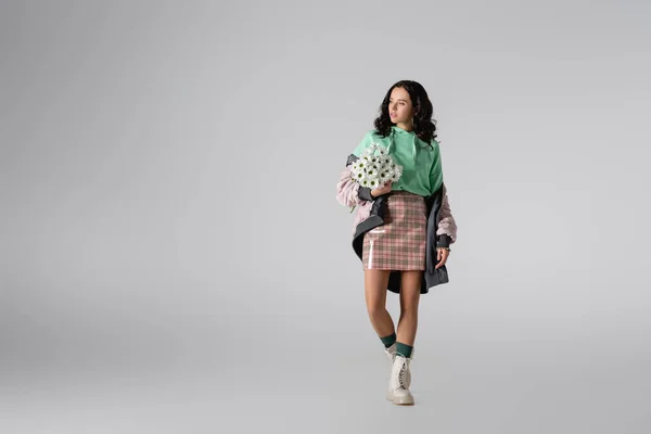 Brunette young woman in stylish winter outfit posing with flowers on grey background — Stock Photo