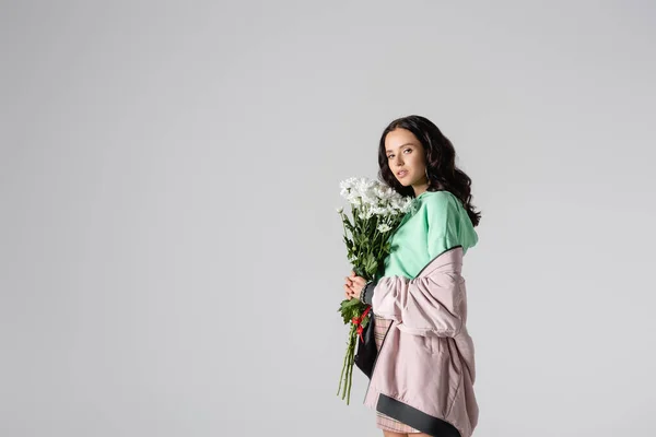 Vista lateral da morena jovem mulher em roupa de inverno elegante posando com flores em fundo cinza — Fotografia de Stock