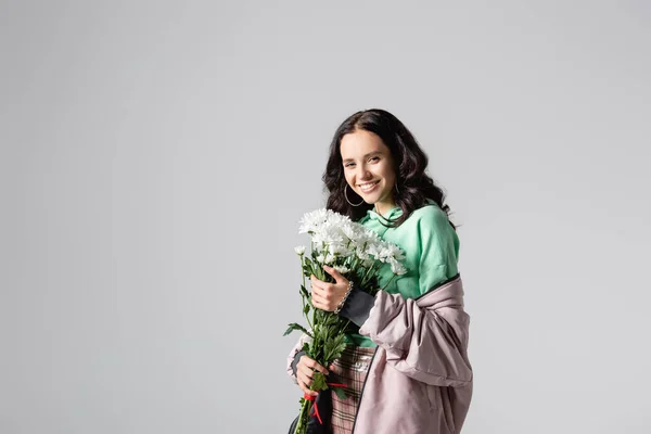 Усміхнена брюнетка молода жінка в стильному зимовому вбранні позує з квітами на сірому фоні — стокове фото