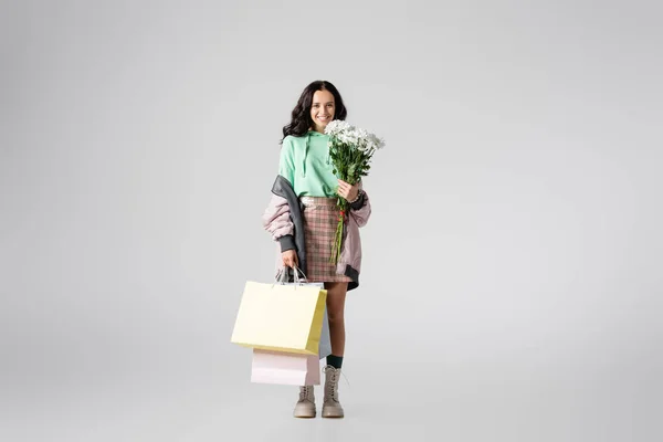 Sorrindo morena jovem mulher em roupa de inverno elegante posando com flores e sacos de compras em fundo cinza — Fotografia de Stock