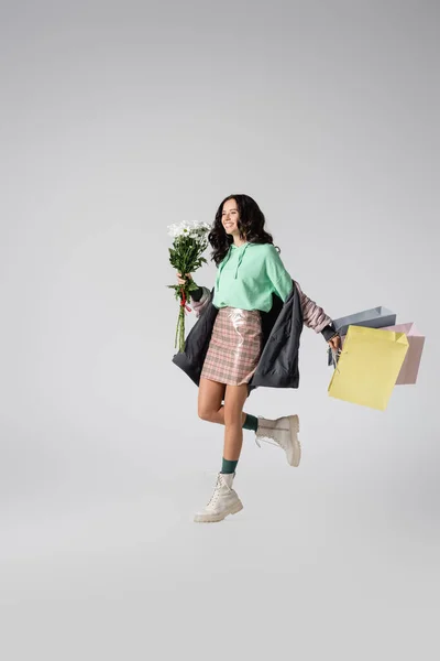 Улыбающаяся брюнетка молодая женщина в стильном зимнем наряде позирует с цветами и сумками для покупок на сером фоне — стоковое фото