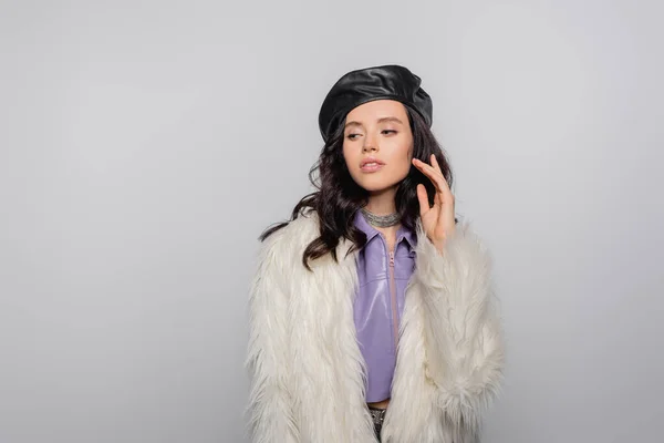 Brune jeune femme en veste blanche élégante en fausse fourrure et béret en cuir posant sur fond gris — Photo de stock