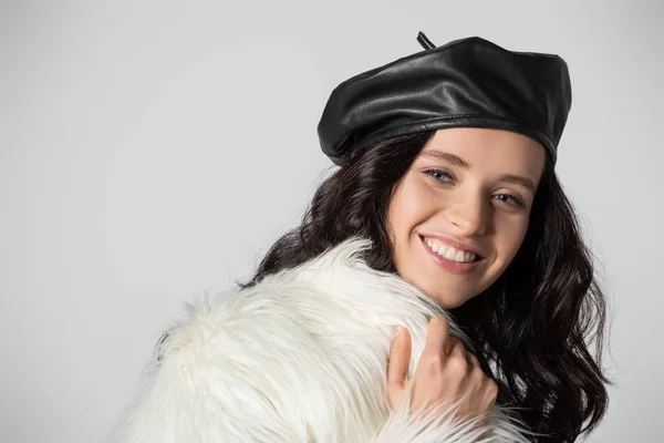 Smiling brunette young woman in stylish white faux fur jacket and leather beret posing on grey background — Stock Photo