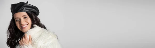 Усміхнена брюнетка молода жінка в стильній білій хутряній куртці та шкіряному береті позує на сірому фоні, банер — стокове фото