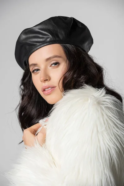 Брюнетка молода жінка в стильній білій хутряній куртці та шкіряному береті позує на сірому фоні — стокове фото