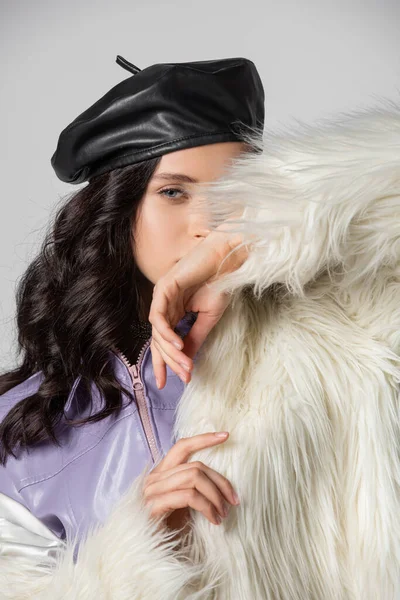 Элегантная брюнетка молодая женщина в стильном белом искусственном меховом пиджаке и кожаном берете позирует на сером фоне — стоковое фото