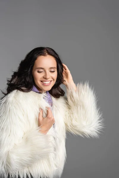 Sonriente elegante morena joven en elegante chaqueta de piel sintética blanca posando sobre fondo gris - foto de stock