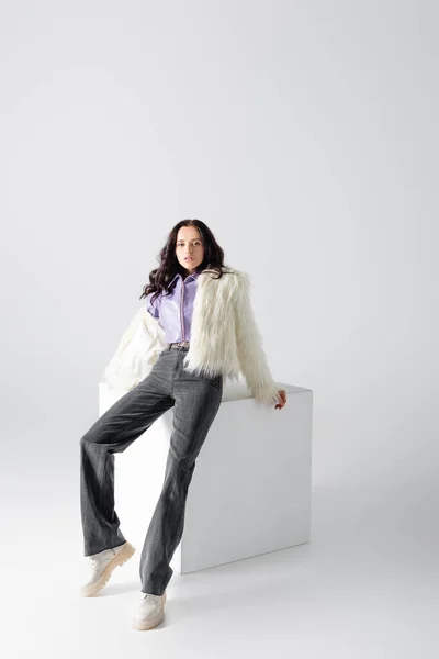 Elegant brunette young woman in stylish faux fur jacket posing on cube on white background — Stock Photo