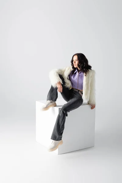 Elegant brunette young woman in stylish faux fur jacket posing on cube on white background — Stock Photo