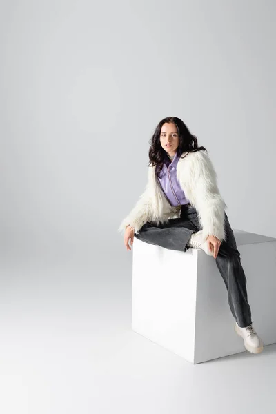 Elegant brunette young woman in stylish faux fur jacket posing on cube on white background — Stock Photo