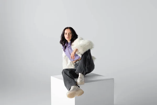 Elegant brunette young woman in stylish faux fur jacket posing on cube on white background — Stock Photo