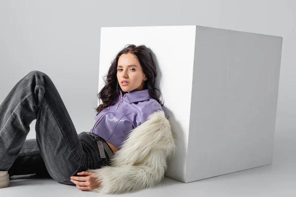 Элегантная брюнетка молодая женщина в стильной искусственной меховой куртке позирует возле куба на белом фоне — стоковое фото