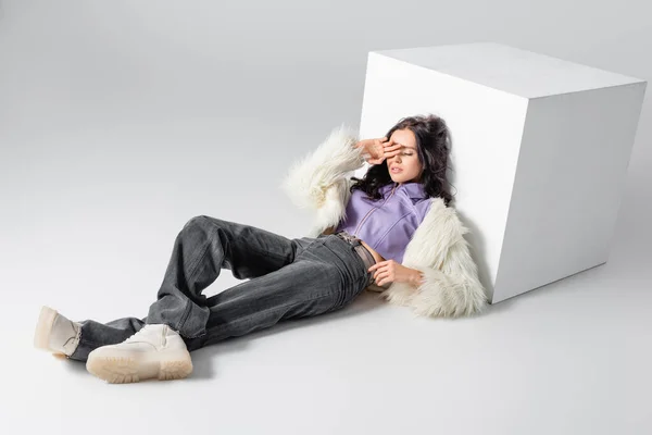 Elegant brunette young woman in stylish faux fur jacket posing near cube on white background — Stock Photo