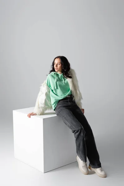 Brunette young woman in faux fur jacket posing near cube on white background — Stock Photo