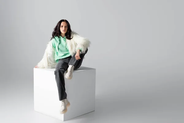 Brunetta giovane donna in pelliccia sintetica giacca in posa sul cubo su sfondo bianco — Foto stock