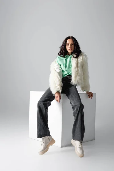 Brunette young woman in faux fur jacket posing on cube on white background — Stock Photo