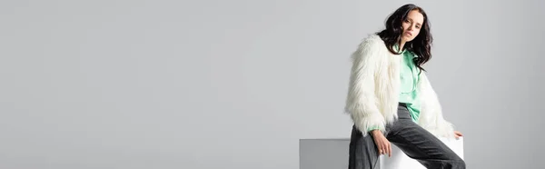 Brunette young woman in faux fur jacket posing on cube on white background, banner — Stock Photo