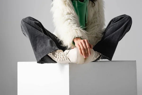 Cropped view of young woman in faux fur jacket and boots posing on cube on white background — Stock Photo