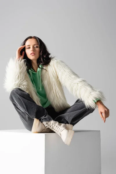 Brunetta giovane donna in pelliccia sintetica giacca in posa sul cubo su sfondo bianco — Foto stock