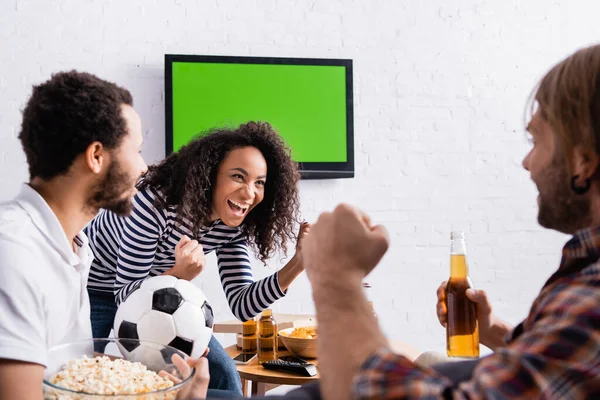 Aufgeregte multiethnische Fußballfans zeigen Siegergeste in der Nähe von lcd tv an Wand auf verschwommenem Vordergrund — Stockfoto