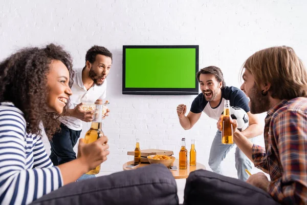 Homem animado com bola de futebol mostrando gesto de vitória perto de amigos multiétnicos segurando cerveja e pipocas — Fotografia de Stock