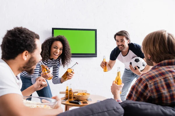 Aficionados al fútbol multiétnicos emocionados celebración de cerveza cerca de lcd tv en la pared en primer plano borrosa - foto de stock